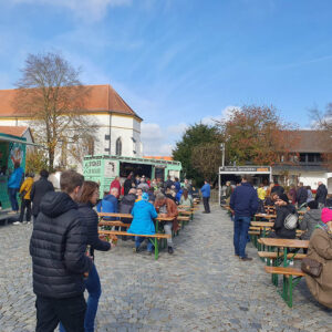 An einem sonnigen Tag sitzen und stehen Menschen an Tischen im Freien in der Nähe von Imbisswagen auf einem Platz mit Kopfsteinpflaster. Im Hintergrund sind eine Kirche und Bäume zu sehen, und der Platz ist voller Besucher, die Essen und Gespräche genießen.
