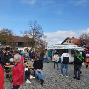 An einem sonnigen Tag versammeln sich Menschen bei einer Veranstaltung im Freien mit Essensständen, Bänken und Luftballons. Einige essen Snacks, stehen oder sitzen auf Bänken auf dem Kopfsteinpflaster. Im Hintergrund sind Bäume und Häuser zu sehen.