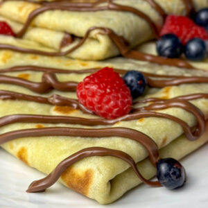 Nahaufnahme von gefalteten Crêpes, die mit Schokoladensauce beträufelt und mit frischen Himbeeren und Blaubeeren garniert sind, serviert auf einem weißen Teller.