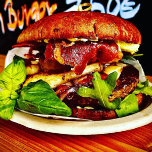 Eine Nahaufnahme eines Gourmet-Burgers auf einem Teller mit knusprigem Speck, gegrilltem Halloumi-Käse, frischem grünen Blattsalat und einer dicken Rindfleischpastete, alles in einem getoasteten Brötchen, mit einem bunten Hintergrund.