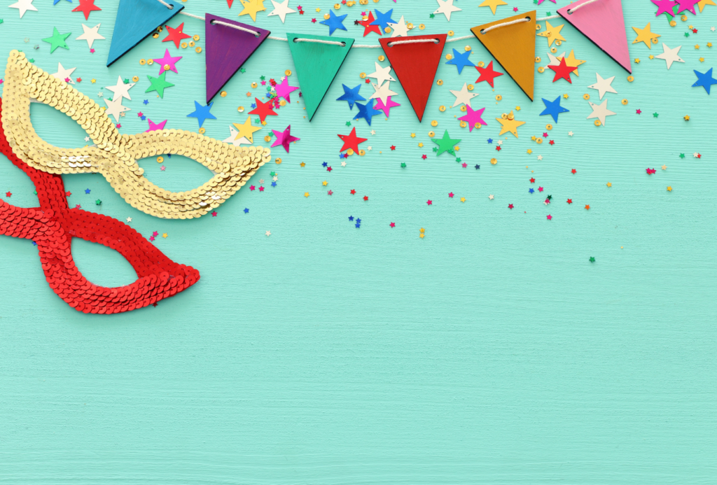Zwei Paillettenmasken in Rot und Gold liegen auf einer türkisfarbenen Fläche, über die buntes, sternförmiges Konfetti verstreut ist. Ein buntes, dreieckiges Wimpelbanner ist darüber drapiert und sorgt für festliche Stimmung.