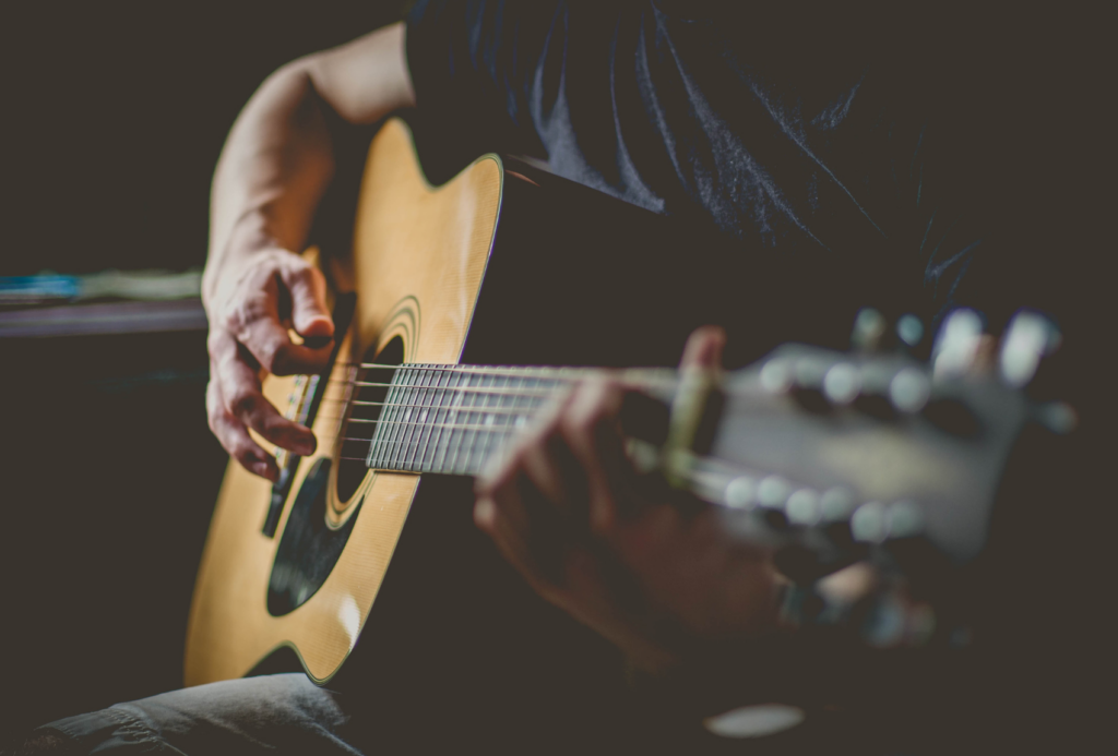 Eine Person spielt Akustikgitarre und konzentriert sich auf ihre Hände, die die Saiten streicheln. Das Foto ist schwach beleuchtet, wodurch das Instrument und die Finger, die sich gekonnt über das Griffbrett bewegen, hervorgehoben werden.