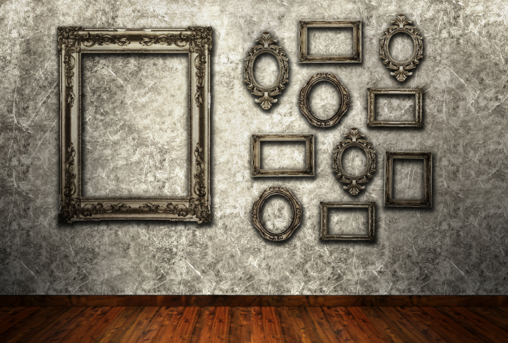 Eine leere Galeriewand mit einer Reihe verzierter Vintage-Bilderrahmen in verschiedenen Formen und Größen auf einer strukturierten, grauen Oberfläche. Ein großer rechteckiger Rahmen steht neben mehreren kleineren Rahmen. Der Raum hat einen Holzboden.
