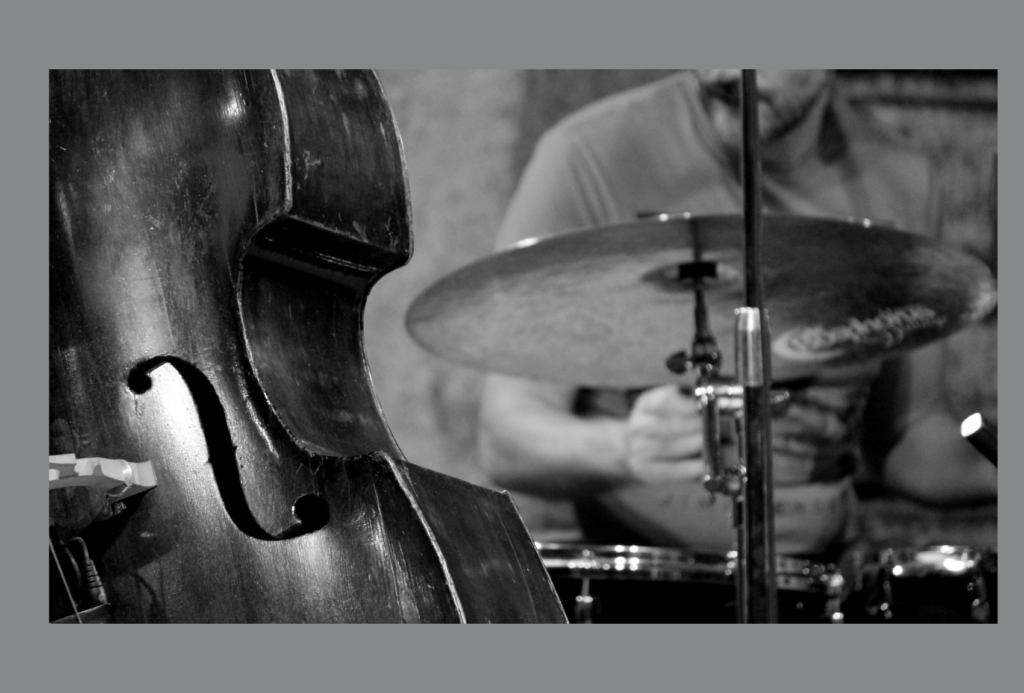 Schwarz-Weiß-Bild eines Schlagzeugspielers im Hintergrund. Im Vordergrund ist ein Kontrabass mit F-Loch und Holzkorpus zu sehen. Der Fokus liegt auf den Instrumenten und vermittelt ein Gefühl von Jazz oder klassischer Musik.