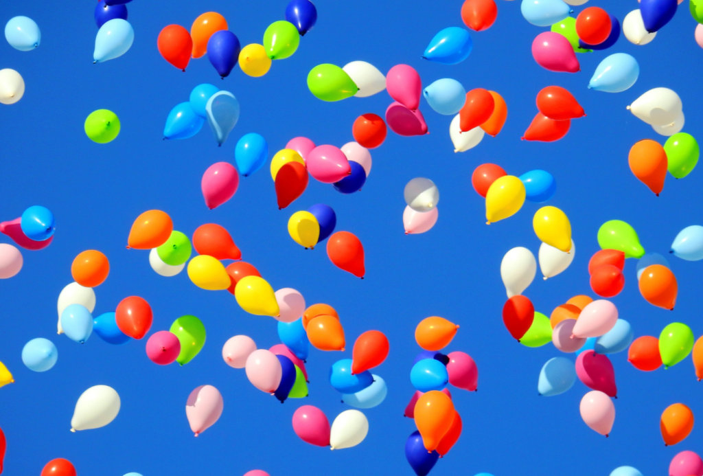 Eine bunte Auswahl an Luftballons in Rot, Blau, Grün, Gelb und Rosa schwebt vor einem strahlend blauen Himmel. Die Ballons variieren in ihrer Größe und sind über das Bild verteilt, wodurch eine fröhliche und festliche Atmosphäre entsteht.