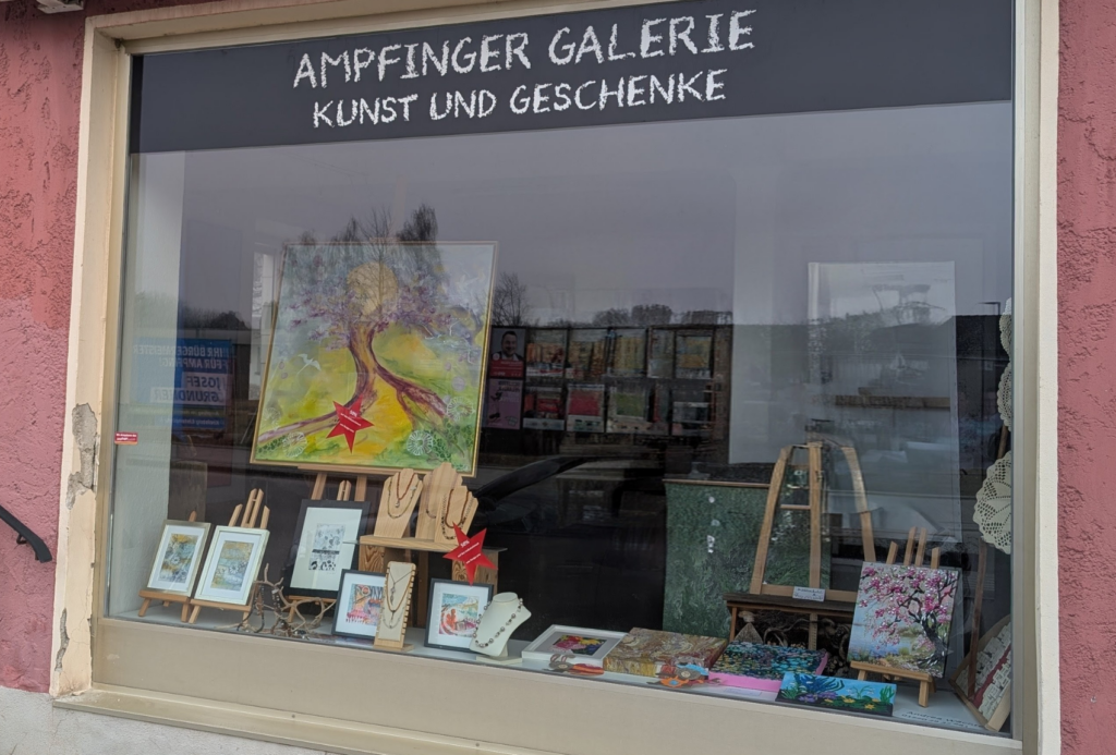 Das Schaufenster der "Ampfinger Galerie Kunst und Geschenke" zeigt Gemälde, gerahmte Kunstwerke, Schmuck, Karten und dekorative Geschenke, die auf Regalen und Staffeleien arrangiert sind, mit Spiegelungen von Bäumen und Gebäuden im Glas.