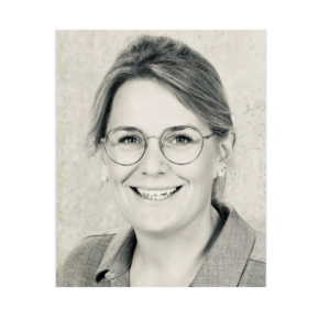 Ein Schwarz-Weiß-Porträt einer lächelnden Person mit mittellangem Haar, Brille und hellem Hemd. Sie blickt direkt in die Kamera vor einem strukturierten Hintergrund.