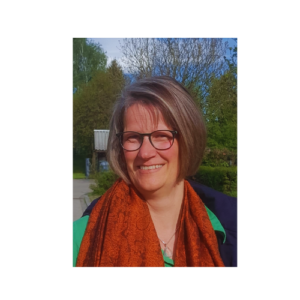 Eine Person mit kurzen grauen Haaren und Brille lächelt und trägt einen orangefarbenen Schal und ein grünes Oberteil. Im Hintergrund sind Bäume und ein klarer blauer Himmel zu sehen.