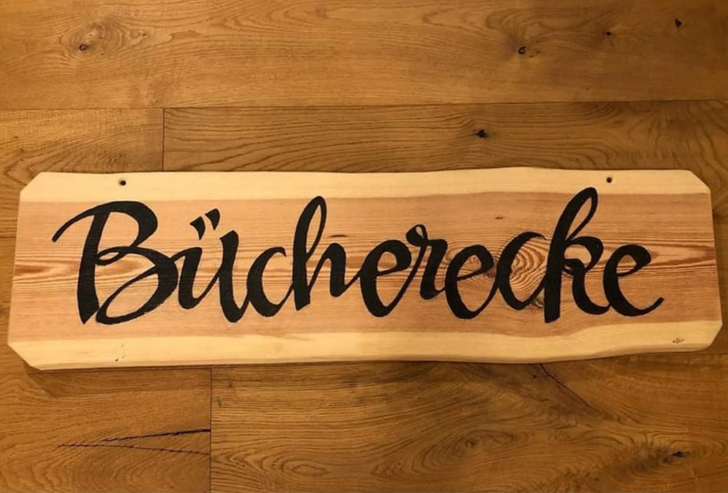 Ein Holzschild mit dem Wort „Bücherecke“ in fetter, schwarzer Schreibschrift auf poliertem Holzhintergrund. Das Schild steht auf einer Holzmaserungsoberfläche.