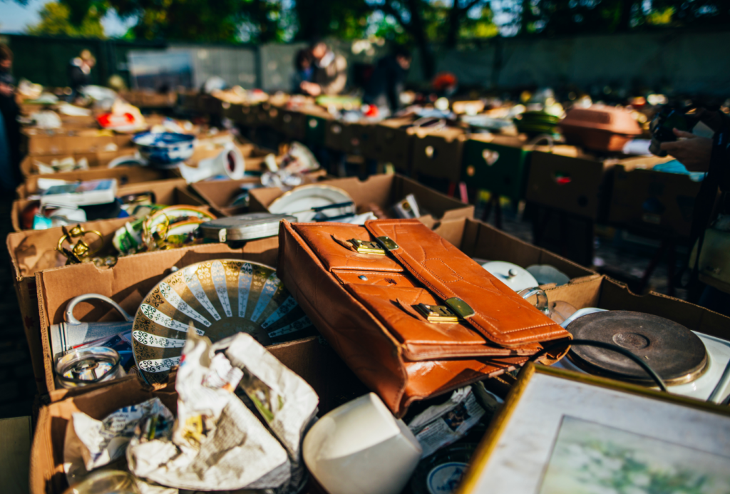 Auf einem Flohmarkt unter freiem Himmel werden auf Tischen im Sonnenlicht Kisten mit verschiedenen Vintage-Artikeln ausgestellt, darunter eine braune Lederaktentasche, Teller, Keramik und gerahmte Bilder.