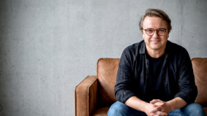 Ein Mann mit Brille, schwarzem Hemd und Jeans sitzt auf einem braunen Ledersofa, lächelt und hat die Hände gefaltet. Der Hintergrund ist eine schlichte graue Wand.