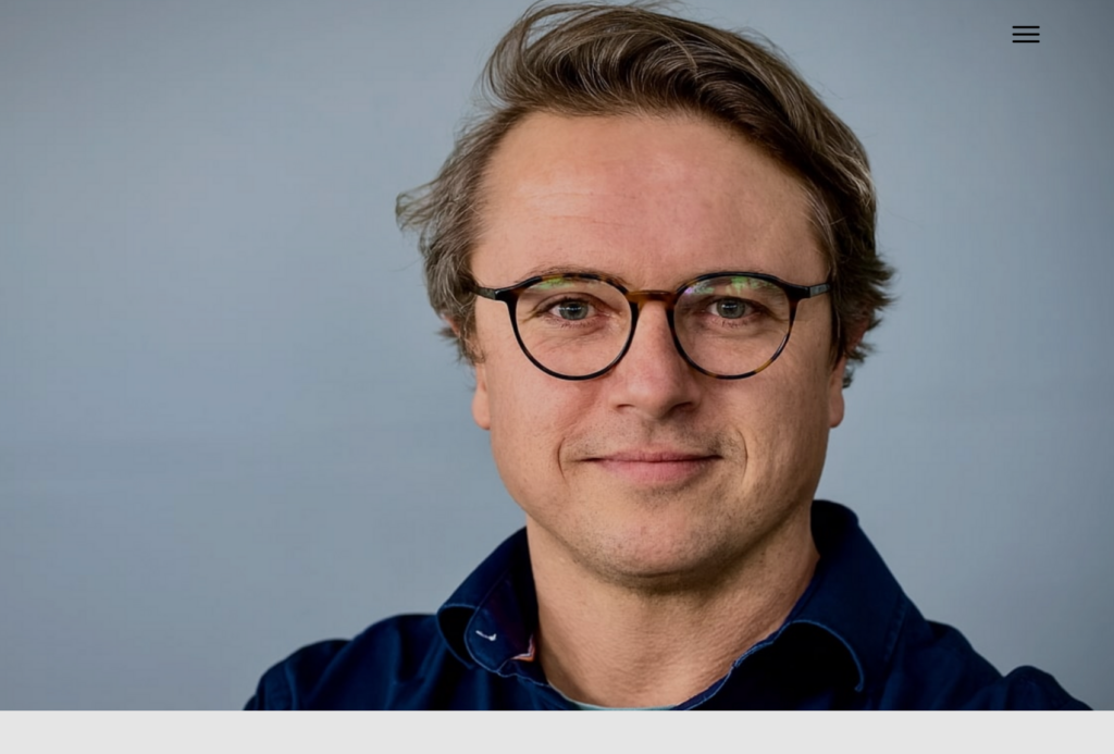 Ein Mann mit hellbraunem Haar, der eine runde Brille und ein dunkelblaues Hemd trägt, schaut mit einem leichten Lächeln direkt in die Kamera. Der Hintergrund ist schlicht und in hellen Farben gehalten.