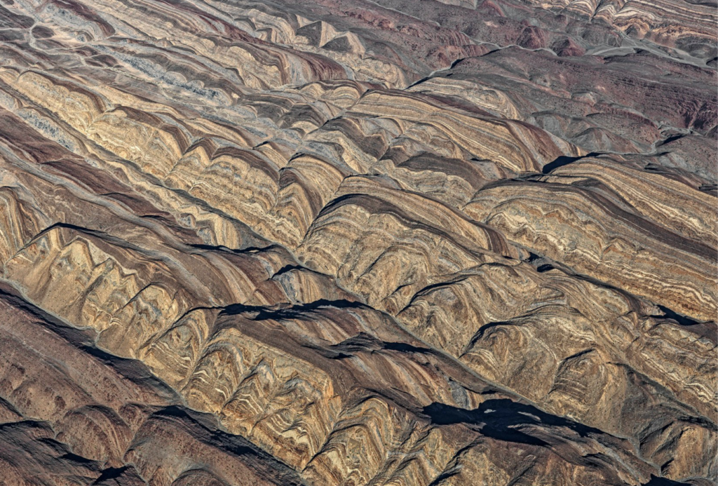Luftaufnahme von geschichteten, gewellten Felsformationen in erdigen Braun-, Hellbraun- und Grautönen, die deutliche geologische Muster und Strukturen in einer zerklüfteten, hügeligen Landschaft zeigen.