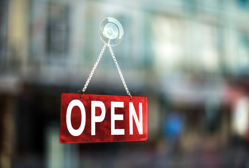 Ein rot-weißes "OPEN"-Schild hängt an einer Kette in einem Schaufenster, mit einer unscharfen Straßenszene im Hintergrund.