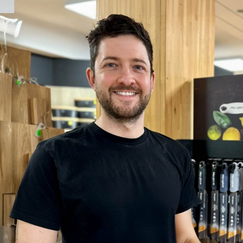 Ein Mann mit kurzen dunklen Haaren und Bart, der ein schwarzes T-Shirt trägt, steht lächelnd in einem Raum vor Holzregalen und Küchenutensilien und hält einen Ampfinger-Gutschein in der Hand.