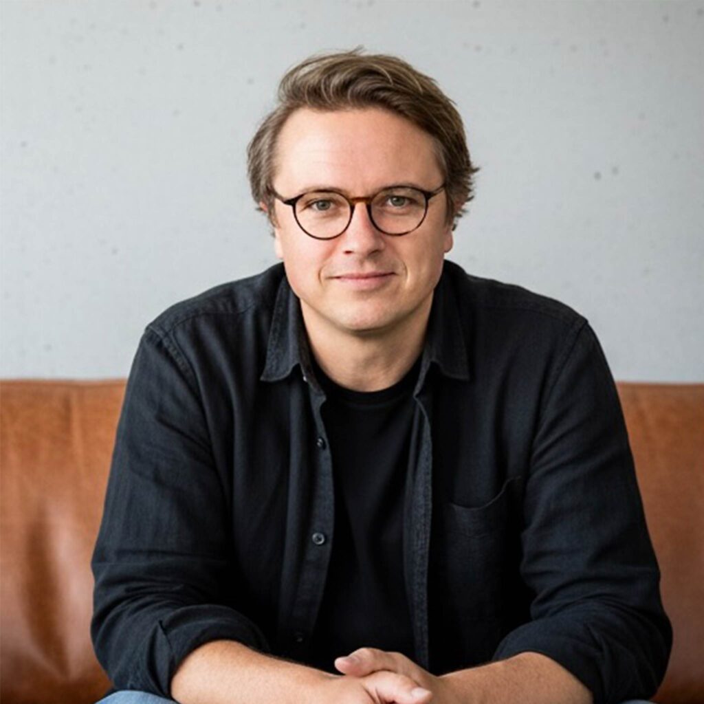 Eine Person mit kurzen, hellbraunen Haaren und Brille, die ein schwarzes Hemd trägt und auf einem braunen Ledersofa sitzt, blickt mit einem leichten Lächeln vor einem schlichten, hellen Hintergrund in die Kamera und hält einen AmPfinger Gutschein in der Hand.