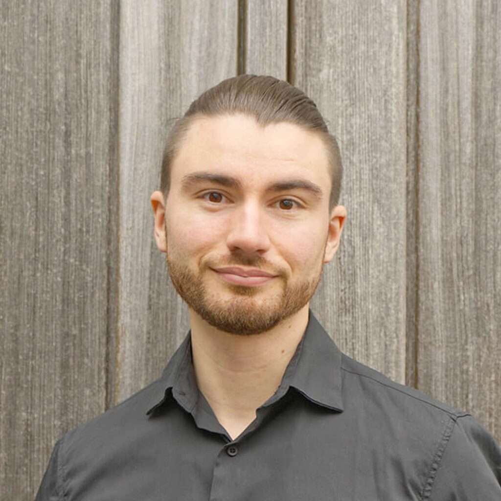 Ein junger Mann mit kurzen braunen Haaren und ordentlich gestutztem Bart, der ein dunkelgraues Button-up-Hemd trägt, steht vor einem Hintergrund mit vertikalen Holztafeln, lächelt leicht in die Kamera und hält einen Ampfinger-Gutschein in der Hand.