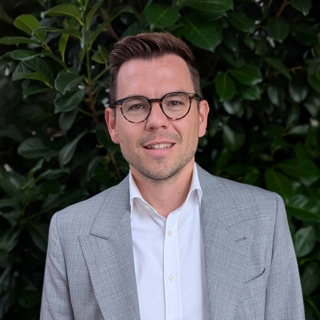 Ein Mann mit Brille, hellgrauer Anzugjacke und weißem Hemd steht vor grünen Büschen, lächelt in die Kamera und hält einen Ampfinger-Gutschein in der Hand.