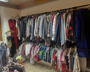 Ein Raum mit zwei parallelen Regalen, die mit verschiedenen Kleidungsstücken für Erwachsene und Kinder gefüllt sind, darunter Jacken, Kleider und Hosen, ordentlich nach Größen geordnet. Ein rotes Kleid hängt separat in der Ecke. Auf dem Boden sind einige Spielsachen zu sehen.