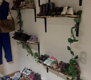 Auf hölzernen Wandregalen mit schwarzen Halterungen stehen Reihen mit verschiedenen Kinderschuhen. Grüne Blätterranken winden sich um die Regale, und an der weißen Wand daneben sind Lichtschalter zu sehen. Auf der linken Seite hängen eine braune Tasche und ein blaues Outfit.