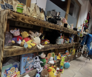 In einem hell erleuchteten Geschäft mit gefliesten Böden und großen Fenstern sind Holzregale mit verschiedenen Kinderspielzeugen, darunter Stofftiere, Puppen, Brettspiele, Puzzles, bunte Bälle und Babyartikel, aufgestellt.