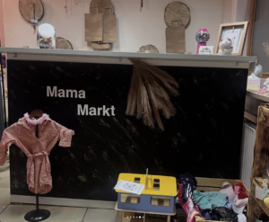 Ein Marktstand mit einer schwarzen Theke mit der Aufschrift "Mama Markt", dekoriert mit getrocknetem Gras. Ein rosafarbenes Baby-Outfit hängt an einem Ständer, und davor sind Kinderspielzeug und -kleidung ausgestellt. Im Hintergrund ist rustikale Wanddekoration zu sehen.