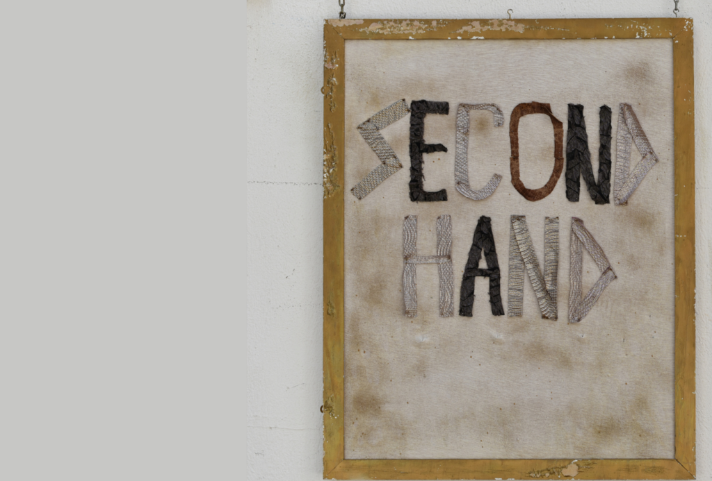 Ein rustikaler Holzrahmen zeigt die Worte "SECOND HAND" in strukturierten, gewebten Buchstaben auf einem verblassten, gebrochen weißen Hintergrund, der an einer weißen Wand hängt.