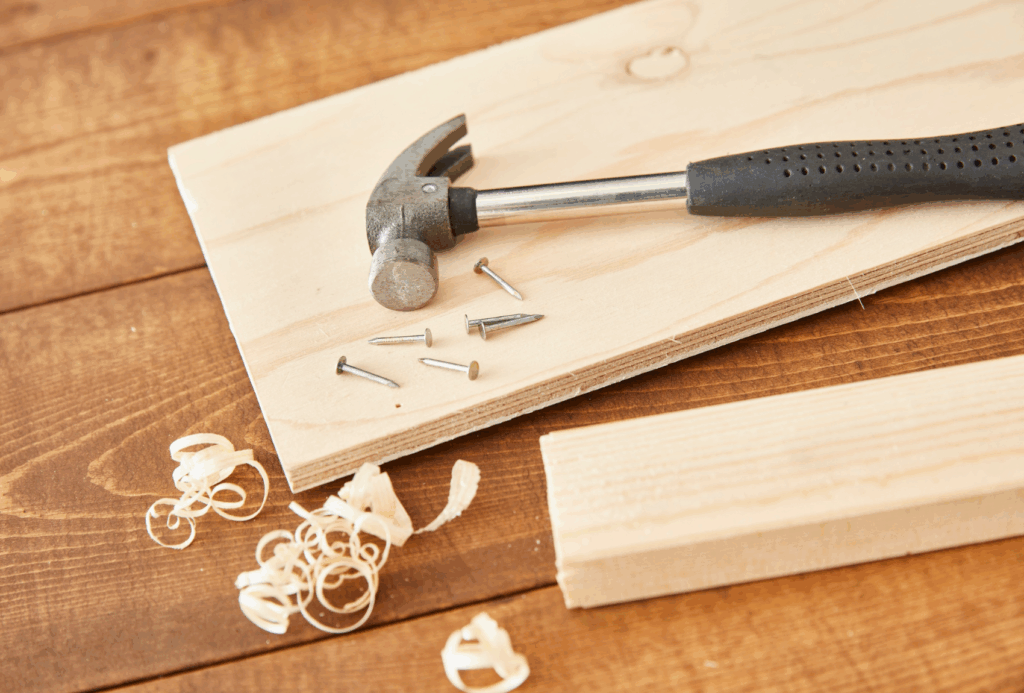Auf einer Holzfläche liegen ein Hammer, Nägel und Holzspäne sowie zwei helle Holzstücke.