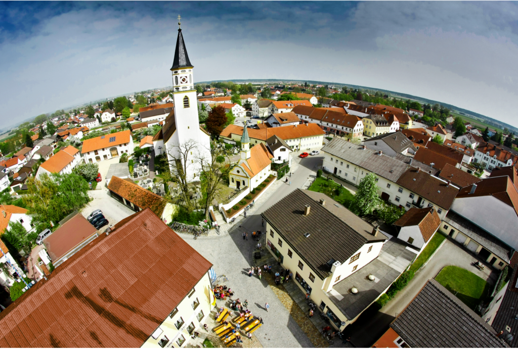 Luftaufnahme einer europäischen Kleinstadt mit einem zentralen Kirchturm, umliegenden Häusern mit roten oder braunen Dächern und Menschen, die sich an einem sonnigen Tag auf einem Platz versammelt haben.