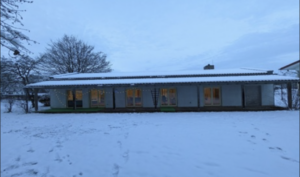 Ein langes, einstöckiges Gebäude mit großen Fenstern ist von Schnee umgeben. Im Inneren leuchten warme Lichter, und im Hintergrund sind kahle Bäume unter einem bewölkten, bläulich schimmernden Himmel zu sehen.