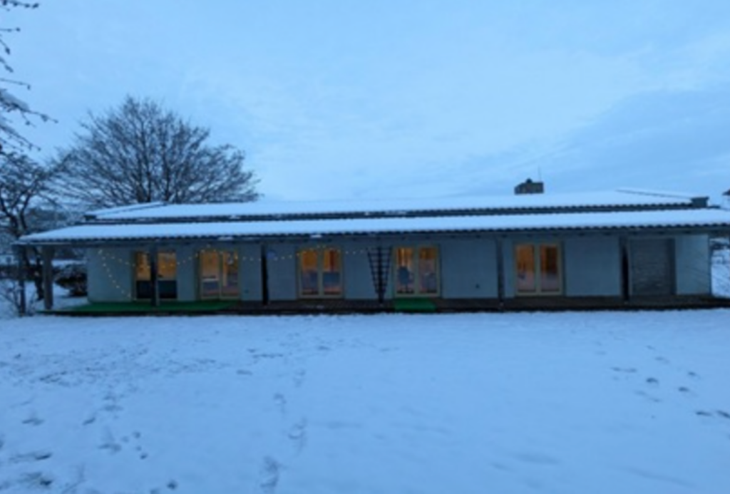 Ein langes, einstöckiges Gebäude mit schneebedecktem Dach steht in der Abenddämmerung in einem verschneiten Garten. Aus den Fenstern und von der Veranda strahlt warmes Licht, und kahle Bäume umgeben das Haus. Im Vordergrund sind Fußspuren im Schnee zu sehen.
