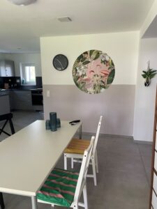 Ein moderner Essbereich mit einem weißen Tisch und zwei Stühlen, von denen einer ein grün gestreiftes Kissen hat. An der Wand befinden sich ein rundes Wandbild mit tropischem Motiv und eine schwarze Uhr. Im Hintergrund sind eine Küche und eine hängende Pflanze zu sehen.