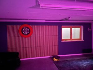 In einem Raum, der in rosa-violettes Licht getaucht ist, befindet sich eine rote Dartscheibe an einer Wand mit vertikalen Vertäfelungen, ein Fenster mit Milchglas sowie ein Sofa, neben dem auf dem Boden ein gelber Gegenstand liegt.