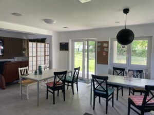 Ein heller Raum mit zwei weißen Tischen und zusammengewürfelten Stühlen, von denen einige mit bunten Kissen ausgestattet sind. Große Fenster und eine Glastür lassen Sonnenlicht herein, und an der Wand hängt eine Pinnwand mit Notizen. Im hinteren Bereich befindet sich eine Sitzecke.