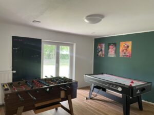 Ein Spielzimmer mit einem Tischfußballtisch, einem Airhockey-Tisch, einer Wandtafel mit Aufschriften und drei bunten Kunstpostern an einer grünen Wand. Große Fenster lassen Tageslicht auf den Holzboden fallen.