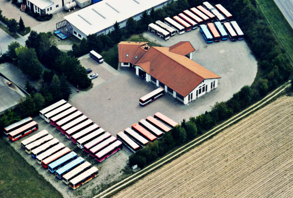 Luftaufnahme eines Gebäudes mit rotem Dach, umgeben von zahlreichen geparkten Bussen, die in Reihen auf einem großen Schotterplatz stehen, der von Bäumen gesäumt ist und an ein Feld grenzt.