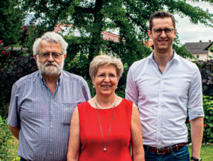 Ein älterer Mann mit weißem Bart, eine ältere Frau in einem roten Kleid und ein jüngerer Mann in einem weißen Hemd stehen gemeinsam im Freien.