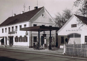 Eine historische Tankstelle mit einem großen Vordach, die zwischen zwei Gebäuden liegt; darauf sind zwei Personen auf Motorrädern zu sehen, die an den Zapfsäulen geparkt haben.