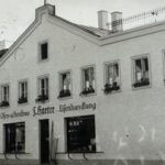 Schwarz-Weiß-Vintage-Foto eines mehrstöckigen Gebäudes mit der Aufschrift „Ofen-u.Herdbau F. Harter Eisenhandlung“ und einer Ladenfront im Erdgeschoss.