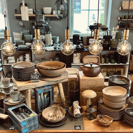 Eine Ausstellung von Geschirr, darunter Stapel von Keramiktellern, Schüsseln, eine Teekanne und verpackte Bestecksets, unter einer Reihe hängender Pendelleuchten.