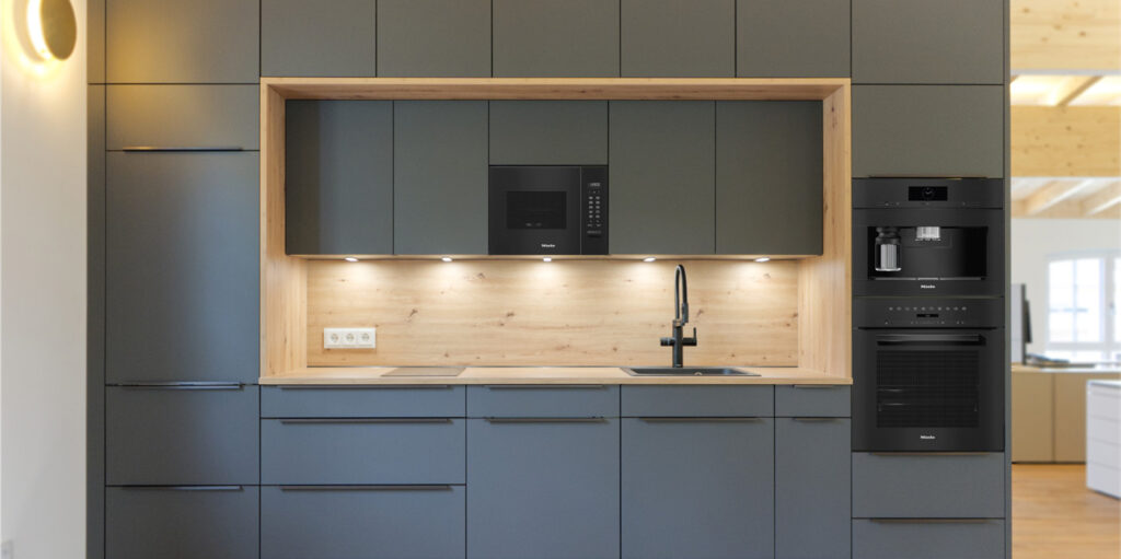 Moderne Küche mit mattgrauen Schränken, einer Küchenrückwand und Arbeitsplatte aus Holz, einer Einbau-Mikrowelle sowie einer schwarzen Miele-Kaffeemaschine und einem Backofen im Stapel.