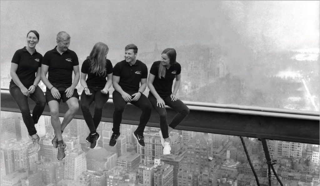 Fünf Menschen in passenden schwarzen Hemden und Hosen sitzen auf einem Stahlträger hoch über einer Stadtlandschaft, die einem berühmten historischen Foto ähnelt. Im Hintergrund ist die Stadt neblig, und die Gruppe wirkt entspannt und lächelnd.