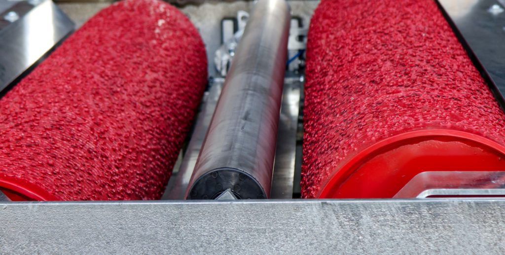 Nahaufnahme einer Industriemaschine mit zwei roten, strukturierten Walzen auf jeder Seite und einer glatten, metallischen Walze in der Mitte. Die Maschine scheint in einem Herstellungsprozess eingesetzt zu werden.