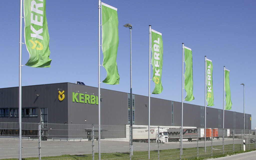 Großes Industriegebäude mit dem „KERBL“-Logo, umgeben von mehreren grünen Flaggen mit demselben Logo. Draußen parken mehrere Sattelschlepper. Der Himmel ist klar und blau. Ein Metallzaun grenzt an den Vordergrund.