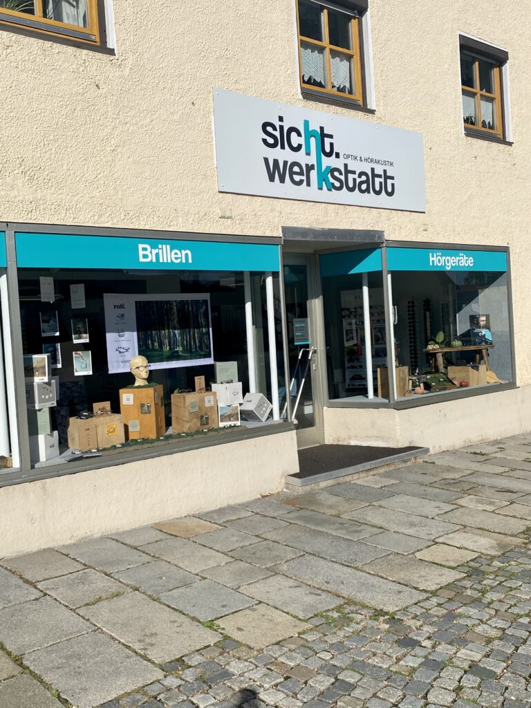 Straßenansicht eines Ladens namens „sicht werkstatt“ mit zwei Schaufenstern. Im linken Schaufenster sind „Brillen“ und verschiedene Schachteln zu sehen, im rechten „Hörgeräte“ mit Hörgerätewerbung. Das Gebäude ist beige mit Kopfsteinpflaster.
