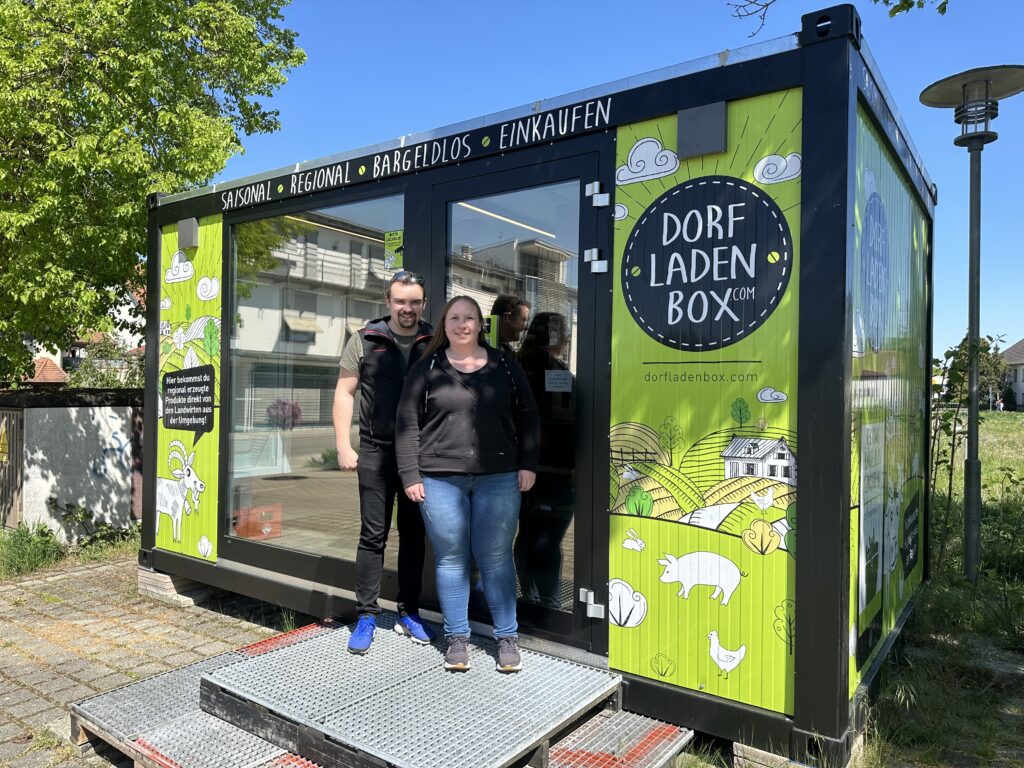 Zwei Personen stehen an einem sonnigen Tag mit Bäumen und Gebäuden im Hintergrund lächelnd vor einem grün-schwarzen Selbstbedienungsladen-Container mit der Aufschrift "Dorfladenbox" und illustrierten lokalen Themen.