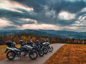 Vier Motorräder sind an einer kurvenreichen Straße geparkt, auf einigen von ihnen liegen Helme. Die Landschaft besteht aus sanften Hügeln und dichten Wäldern unter einem dramatischen, wolkigen Himmel, der darauf schließen lässt, dass ein Sturm aufzieht. Die Szene vermittelt ein Gefühl von Abenteuer.