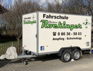 Ein mobiler Anhänger mit der Aufschrift „Fahrschule Rockinger“ und Kontaktinformationen, inklusive Telefonnummer und Standorten (Ampfing – Schwindegg) in grüner und schwarzer Schrift. An der Seite ist ein QR-Code sowie verschiedene Logos sichtbar.