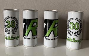 Vier hohe Dosen mit einem Design, bei dem auf zwei Dosen die Zahl „30“ in einem Lorbeerkranz und auf den anderen beiden die Buchstaben „FR“ zu sehen sind. Darunter ist auf jeder Dose in Grün und Schwarz das Wort „Fahrschule“ aufgedruckt. Sie stehen auf einer weißen Fläche.