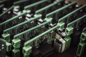 Nahaufnahme mehrerer grüner Leiterplatten, die in einer Reihe angeordnet sind. Die Platten sind vertikal ausgerichtet und dicht gepackt und zeigen verschiedene elektronische Komponenten wie Widerstände und Anschlüsse in einer Fertigungs- oder Montageumgebung.